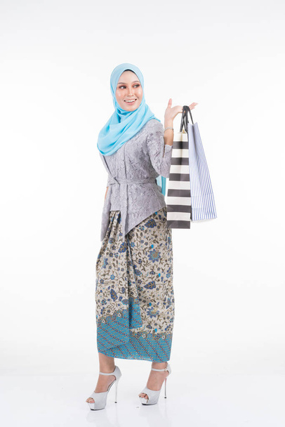A beautiful Muslim female model in a Asian traditional dress modern kebaya and hijab carrying shopping bags isolated on white background. Eidul fitri fashion and festive shopping concept - Fotografie, Obrázek