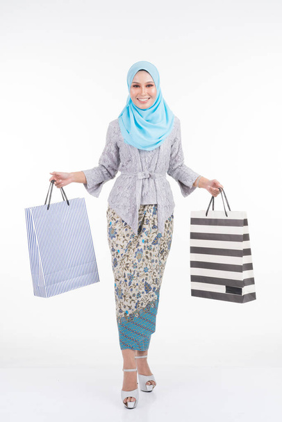 A beautiful Muslim female model in a Asian traditional dress modern kebaya and hijab carrying shopping bags isolated on white background. Eidul fitri fashion and festive shopping concept - Foto, Bild