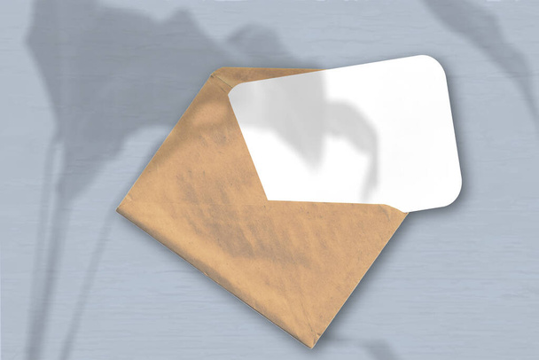 Ein Umschlag mit einem Blatt strukturiertem weißem Papier auf grauem Hintergrund. Mockup mit einer Überlagerung von Pflanzenschatten. Natürliches Licht wirft Schatten von einer exotischen Pflanze. Horizontale Orientierung. - Foto, Bild