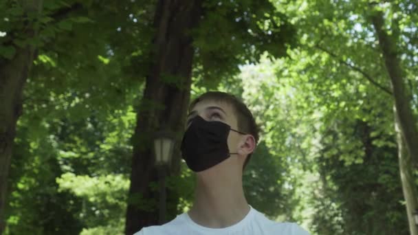 Νεαρός άνδρας με ιατρική προστατευτική μαύρη μάσκα σε δάσος με πράσινο φύλλωμα και δέντρα σε ηλιόλουστη μέρα. Ταξιδιωτική ιδέα μετά την επιδημία του Coronavirus. Άνοιγμα των συνόρων και έναρξη της τουριστικής περιόδου - Πλάνα, βίντεο
