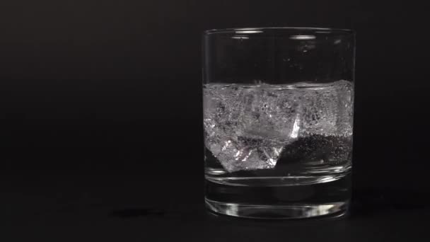 Close-up of carbonated transparent clear water mixed in a glass with ice cubes on a black background. Slow motion. Bubble swirl - Video, Çekim