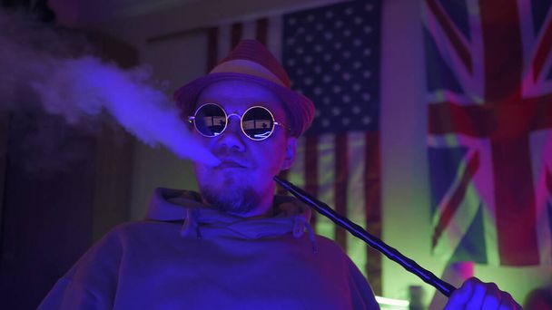 Creative man relaxes at home in evening. Young hipster smokes hookah exhales cloud of smoke at side while looking at camera. Against background of of USA and UK flags hanging on wall in dark room - Photo, image