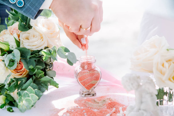 Bride and groom pouring colorful different colored sands into the crystal vase close up during symbolic nautical decor destination wedding marriage ceremony on sandy beach in front of the ocean  - Foto, afbeelding