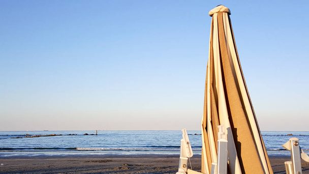 Parasole i leżaki zamknięte o świcie nad brzegiem morza, z parasolami i leżakami zamkniętymi o świcie nad brzegiem morza, ze słońcem odbijającym się od wiatraków. Wysokiej jakości zdjęcie - Zdjęcie, obraz