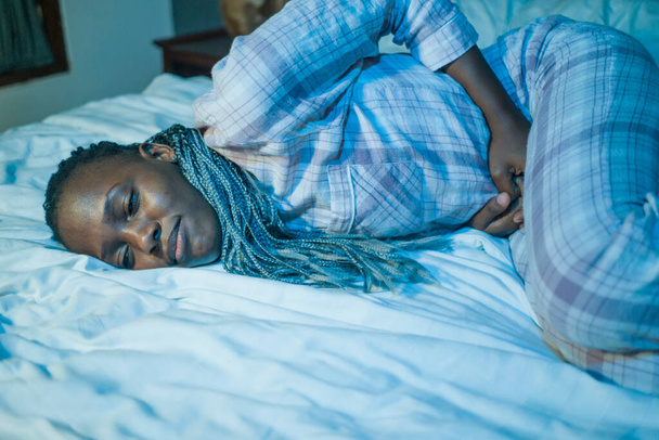 drámai otthoni portré fiatal beteg és depressziós afro amerikai nő pizsamában feküdt az ágyban szenvedő menstruáció és a menstruációs fájdalom rossz közérzet tartja a gyomor álmatlan éjszaka - Fotó, kép