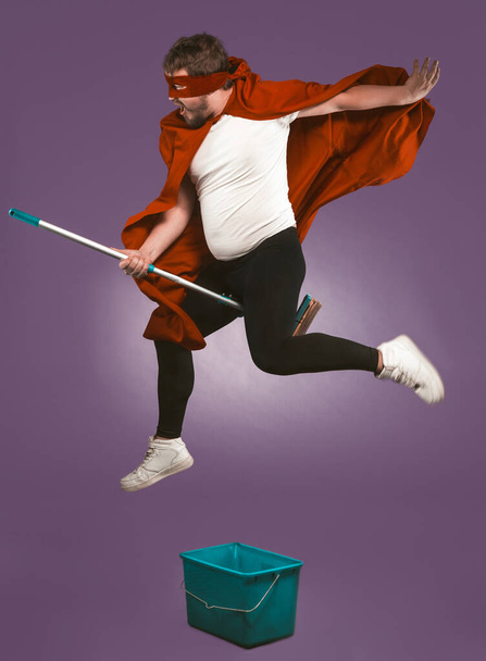 Super hero flying on mop. Funny man have fun with a bucket and a mop on grape purple background. Funny cleaning concept - Foto, immagini