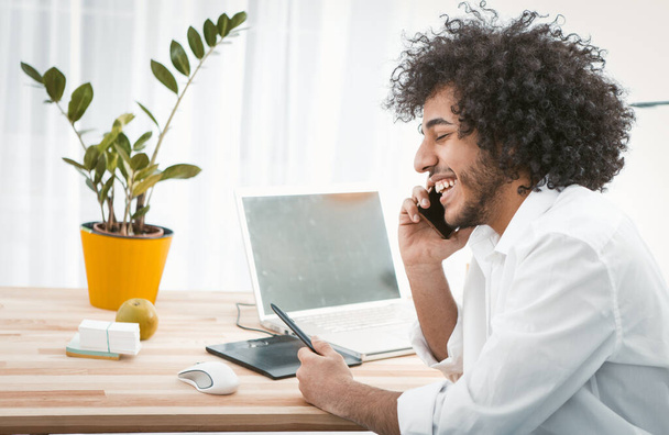 Creative man communicates by mobile phone smiling while sitting at home workplace. Freelance concept. Work from home concept. Side view. Copy space at left side. Toned image - Photo, image