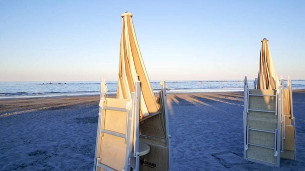 Parasole i leżaki zamknięte o świcie nad brzegiem morza, z parasolami i leżakami zamkniętymi o świcie nad brzegiem morza, ze słońcem odbijającym się od wiatraków. Wysokiej jakości zdjęcie - Zdjęcie, obraz