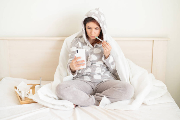 Woman sitting on the bed covered with a blanket and photographed on the phone with a thermometer in his mouth. A girl with a cold takes a selfie on her blog. Measurement of elevated temperature. - 写真・画像