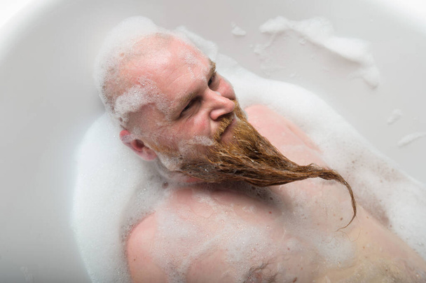 A bald man is fooling around in the foam bath. The guy makes funny hairstyles on his red beard. - Фото, зображення