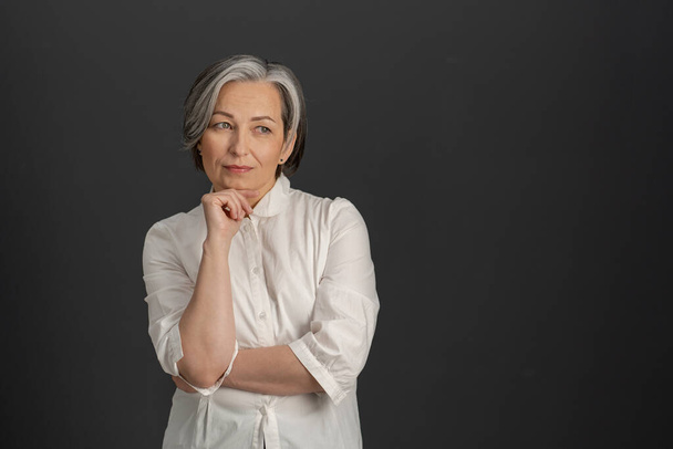 Thoughtful mature woman thinks touching by hand of her chin while looking at side. Gray-haired lady in white shirt isolated on gray background. Textspace at right side - Foto, immagini