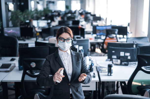 Female manager in a medical mask sprays on hand antiseptic. A woman in a suit uses a sanitizer to disinfect. Concept of office work during the coronavirus epidemic. - Foto, Bild