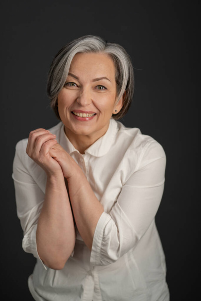 Joyful Caucasian woman smiles broadly while looking at camera and clasped her hands. Enthusiastic gray-haired mature woman in a white shirt posing in studio - Foto, afbeelding