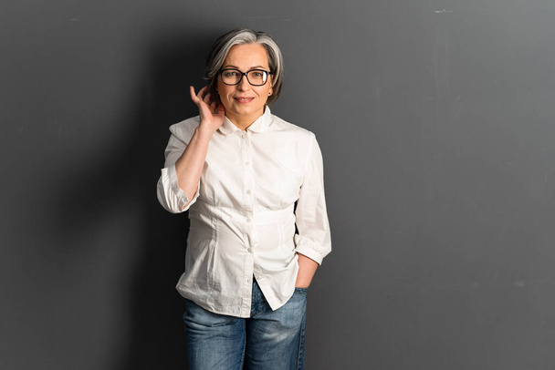 Pretty mature woman corrects medium-length hairstyle coquettishly. Adorable Gray-haired Caucasian model in jeans posing on gray background. Copy space or textspace at right side - Fotografie, Obrázek