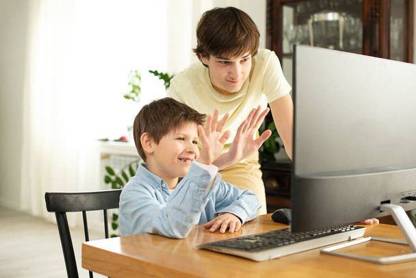 Garoto sorridente e adolescente conversando online e acenando para a tela do computador. Quarentena e auto-isolamento devido ao coronavírus. COVID-19
 - Foto, Imagem