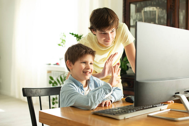 Garoto sorridente e adolescente conversando online e acenando para a tela do computador. Quarentena e auto-isolamento devido ao coronavírus. COVID-19
 - Foto, Imagem