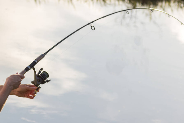 Το καλάμι ψαρέματος στα χέρια ενός ψαρά στο ποτάμι. όμορφα χέρια με κλώση στο φόντο του νερού. έννοια για την ημέρα του ψαρά και το καλοκαίρι αλιείας. χόμπι - Φωτογραφία, εικόνα
