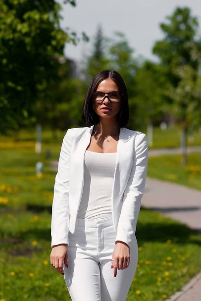 Confident sensual woman in stylish white suit standing with eyes closed and arms raised and enjoying life and freedom on street against blurred walkway and green trees growing on flowered lawns - Photo, Image