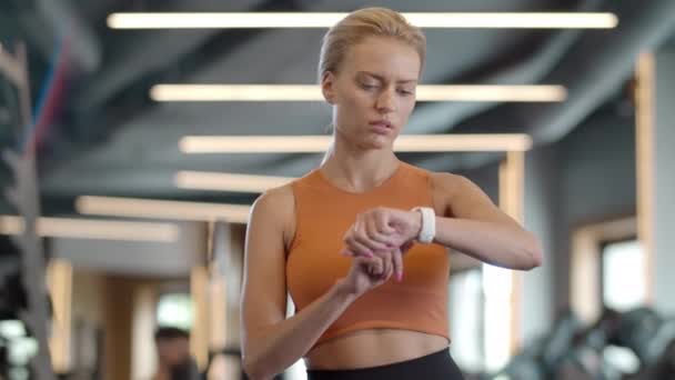 Aufgeregt blickt eine Frau im Fitnessstudio auf die Armbanduhr. Sportlerin steht im Sportverein - Filmmaterial, Video