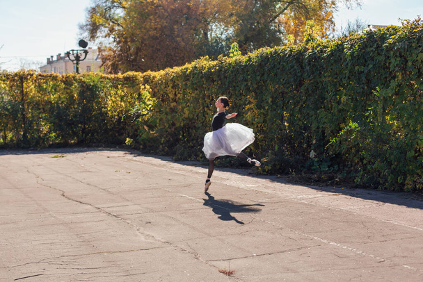 Woman ballerina in a white ballet skirt dancing in pointe shoes in a golden autumn park. Ballerina jumping doing a split - 写真・画像