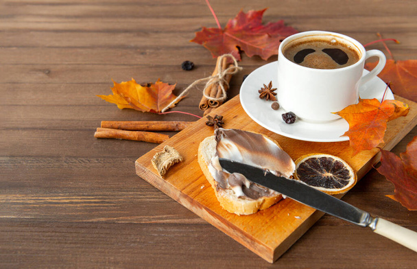 Cup of black coffee with a saucer, orange autumn maple leaves, cinnamon sticks, anise stars ,a piece of bread with creamy chocolate paste on a brown wooden background - Fotografie, Obrázek