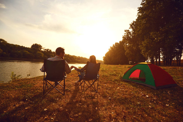 A hiker couple holding hands next to a tent by the lake at sunset. - 写真・画像