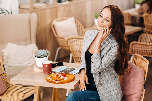A girl is sitting in a cafe and talking on the phone, a Girl in a coffee shop is smiling and burning on a smartphone, sweets are on the table - Foto, Imagen