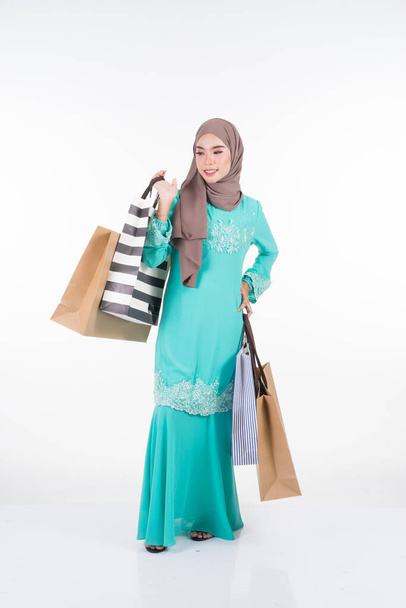 A beautiful and excited Muslim female model in a Asian traditional dress modern kurung carrying shopping bags isolated on white background. Eidul fitri festive preparation shopping concept. Full length portrait. - Foto, Imagem