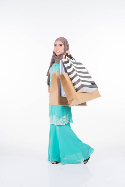Ένα όμορφο και ενθουσιασμένο μουσουλμανικό θηλυκό μοντέλο σε ένα ασιατικό παραδοσιακό φόρεμα σύγχρονο kurung μεταφέρουν τσάντες ψώνια που απομονώνονται σε λευκό φόντο. Eidul fitri εορταστική προετοιμασία έννοια ψώνια. Πλήρες μήκος πορτρέτο. - Φωτογραφία, εικόνα