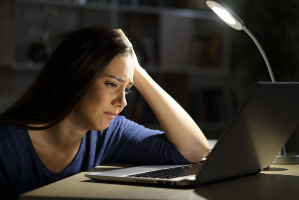 Sad woman looking at laptop complaining sitting alone at night at home - Фото, изображение