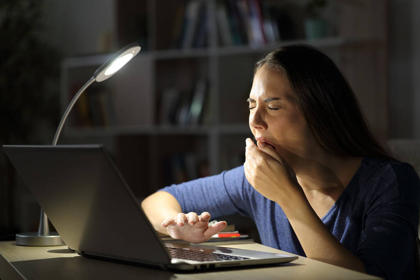 Tired woman with laptop yawning sitting late with a desk lamp on the table at night at home - Foto, immagini
