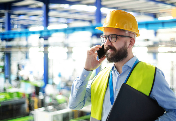 Technician or engineer with hard hat standing in industrial factory, using telephone. - 写真・画像