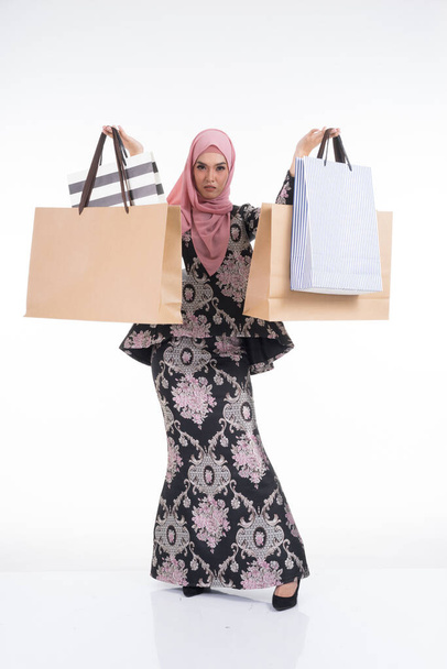 Ένα όμορφο ασιατικό θηλυκό μοντέλο σε ένα παραδοσιακό φόρεμα που μεταφέρουν τσάντες ψώνια που απομονώνονται σε λευκό φόντο. Eidul fitri εορταστική προετοιμασία έννοια ψώνια. Πλήρες μήκος πορτρέτο. - Φωτογραφία, εικόνα