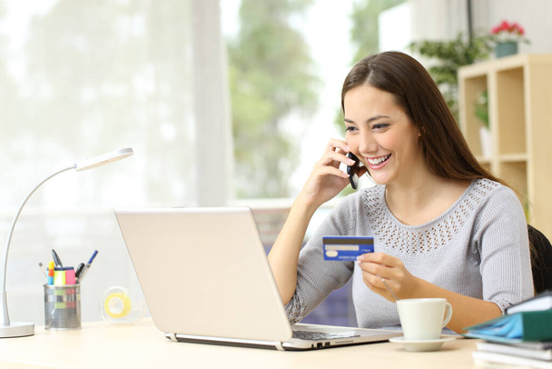 Happy woman with laptop talking on smart phone holding credit card sitting on a desk at home - Foto, immagini
