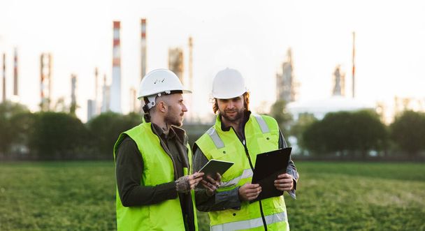 Two young engineers with tablet standing outdoors by oil refinery, discussing issues. - Foto, Imagen