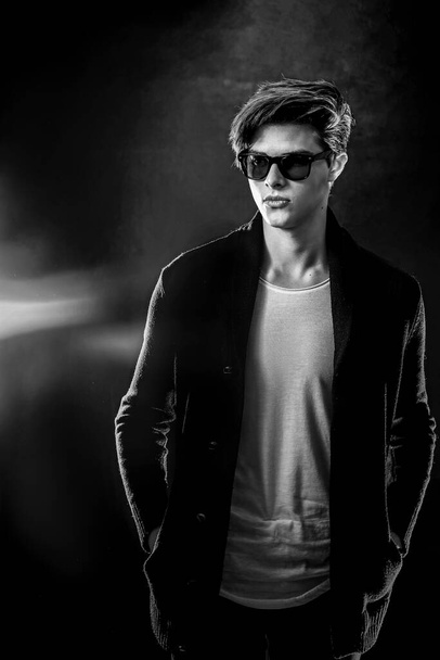 Cool stylish man in black jacket and sunglasses. High Fashion male model posing on black background. - Фото, изображение
