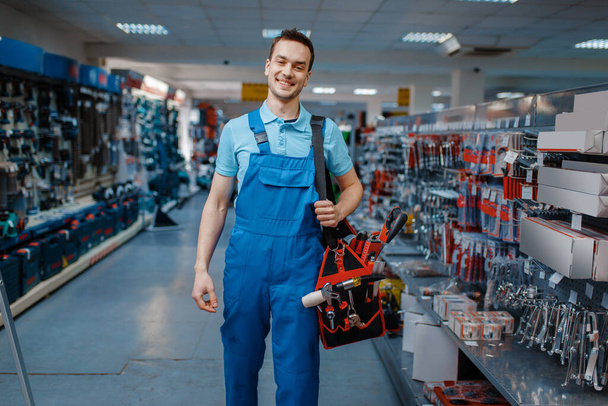Üniformalı erkek işçi alet çantasını alet deposunda tutuyor. Hırdavatçıda profesyonel ekipman seçimi - Fotoğraf, Görsel