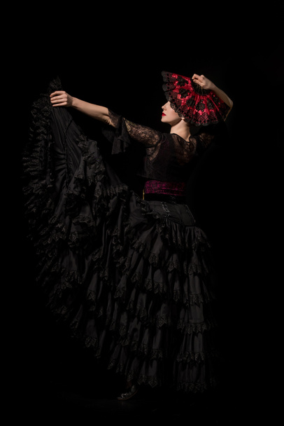 young and beautiful dancer holding fan while dancing flamenco isolated on black  - Foto, afbeelding