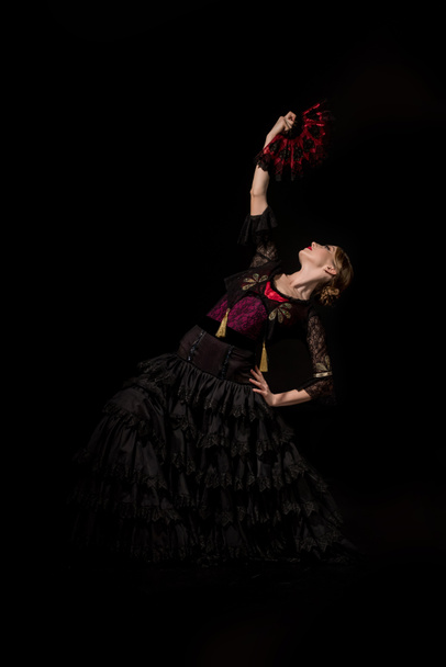 pretty flamenco dancer with hand on hip holding fan and dancing isolated on black  - Valokuva, kuva