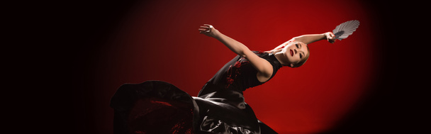 panoramic shot of young flamenco dancer in dress holding fan while dancing on red  - Fotografie, Obrázek