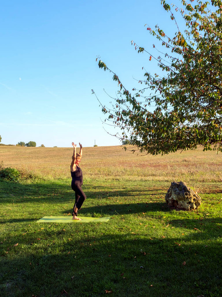 Surrounded by idyllic rural French countryside a mature woman practises her yoga positions in beautiful autumn sunshine. The Tree pose. - Photo, Image