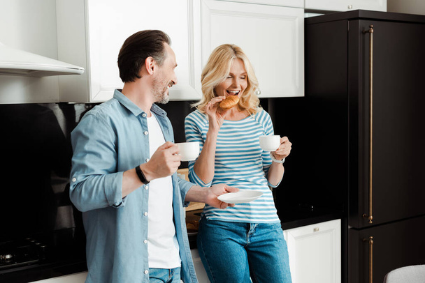 Lächelnder Mann mit Kaffeetasse und Blick auf Frau, die in Küche Croissant isst  - Foto, Bild