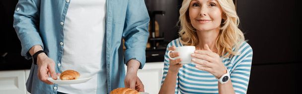 Panoramic crop of smiling woman holding cup of coffee near husband holding croissants in kitchen  - Photo, Image
