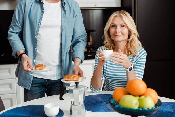 Enfoque selectivo de la mujer sonriente con taza de café cerca del hombre con croissants en la cocina
  - Foto, Imagen