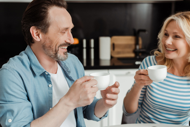 Beautiful smiling woman holding cup of coffee near husband in kitchen  - Photo, Image