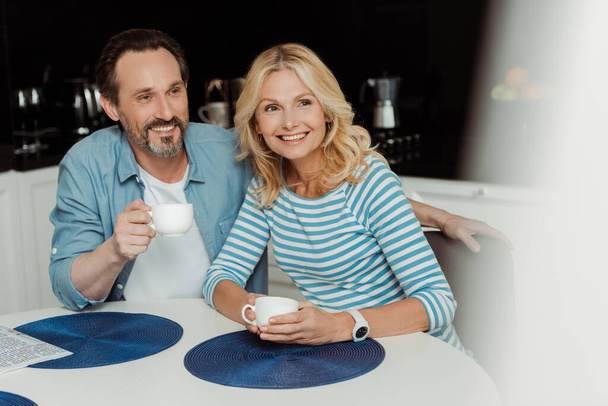 キッチンでコーヒーを飲みながら笑顔の正の成熟したカップルの選択的な焦点  - 写真・画像