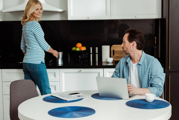 Smiling woman holding geyser coffee maker and smiling at husband using laptop in kitchen  - Фото, зображення