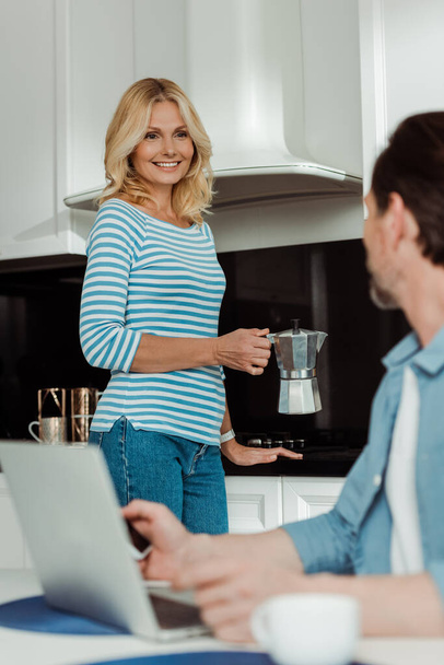 Enfoque selectivo de la mujer sosteniendo la cafetera géiser y sonriendo al marido usando aparatos en la cocina
  - Foto, imagen