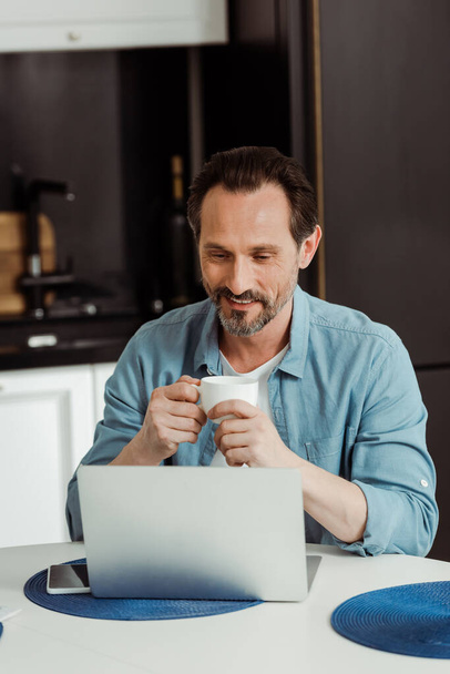 Selective focus of smiling mature man using laptop and drinking coffee in kitchen  - Photo, Image