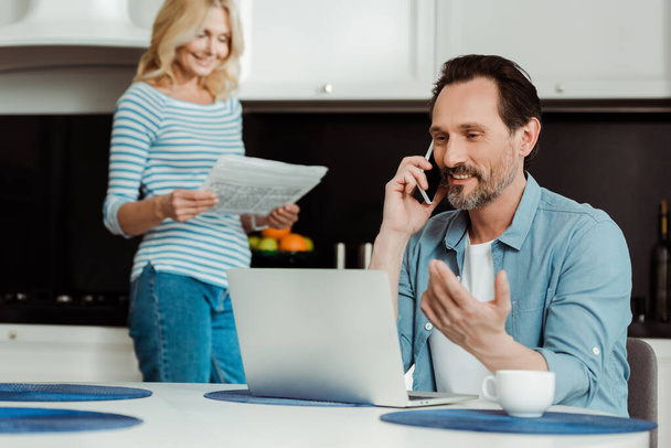 Enfoque selectivo del hombre sonriente hablando en un teléfono inteligente cerca de la computadora portátil mientras su esposa lee el periódico en la cocina
  - Foto, imagen